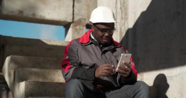 Afrikalı Amerikalı müteahhit inşaat alanında merdivenlerde oturur ve video bağlantısıyla akıllı telefondan konuşur. Sinema 4K 60fps video