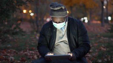 Tıbbi maskeli Afrikalı Amerikalı adam sonbahar akşamı parkında oturup tablet bilgisayarında oyun oynuyor.