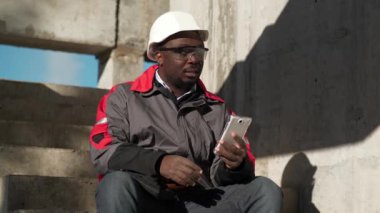 Slow Motion Afrikalı Amerikalı inşaatçı inşaat alanındaki merdivenlerde oturup video bağlantısıyla akıllı telefondan konuşuyor.
