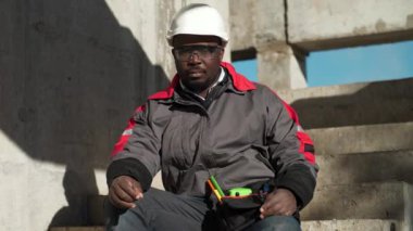 Afrikalı Amerikalı işçi inşaat alanındaki merdivenlerde oturur ve kameraya bakar.
