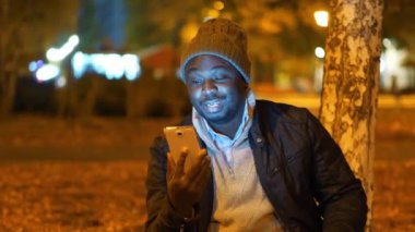 Kış şapkalı Afrikalı Amerikalı adam akşam parkında oturuyor ve akıllı telefondan video bağlantısıyla konuşuyor. Yapay ışıklandırmanın açık hava lambalarından titreyen video