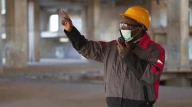 Siyah şapkalı, gözlüklü ve tıbbi maskeli Afrikalı Amerikalı müteahhit inşaat alanında duruyor ve taşınabilir radyo vericisiyle konuşuyor.