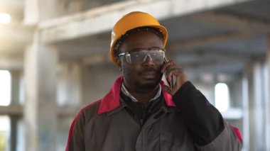 Slow Motion Afrikalı Amerikalı işçi inşaat alanında akıllı telefondan konuşuyor. Sarı başlıklı işçi cep telefonundan konuşuyor.