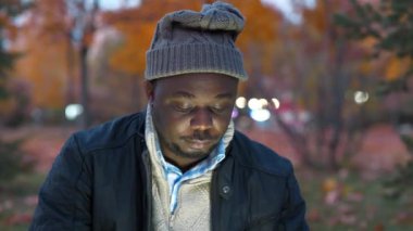 Kış şapkalı Afrikalı Amerikalı adam sonbahar akşamı parkında oturur ve tablet bilgisayarının parlak ekranına bakar, doğada çalışır.