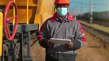 Elinde tablet bilgisayarla tıbbi maske takan Afro-Amerikan demiryolu çalışanı yük treni terminalinde kameraya bakıyor. Yük istasyonunda tabletli demiryolu trafiği müfettişi.
