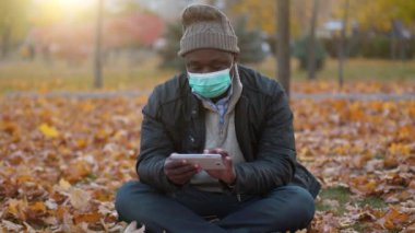 Kış şapkalı ve tıbbi koruyucu maskeli Afrikalı bir adam sonbahar güneşli parklarda sarı yapraklara oturur ve akıllı telefonuyla çalışır.