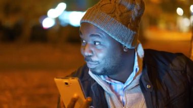 Kış şapkalı Afrikalı Amerikalı bir adam akşam parkında oturuyor ve akıllı telefondan video bağlantısıyla konuşuyor.