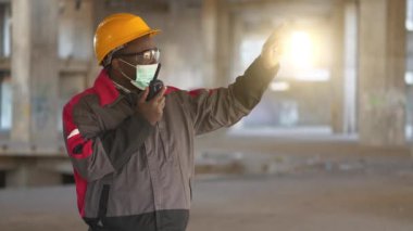 Sarı şapka, gözlük ve tıbbi maske takan Afrikalı Amerikalı müteahhit inşaat alanında duruyor ve taşınabilir radyo vericisiyle konuşuyor.