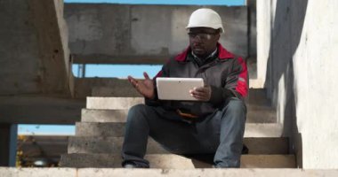 Beyaz şapkalı yorgun Afrikalı Amerikalı işçi inşaat alanındaki merdivenlerde oturup tablet bilgisayarla video bağlantısıyla konuşuyor. Sinema 4K 60fps video