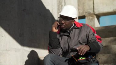 Slow Motion Afro-Amerikan çalışanı akıllı telefondan konuşuyor. Beyaz şapkalı ve koruyucu gözlüklü inşaatçı inşaat alanında merdivenlerde oturuyor ve cep telefonuyla konuşuyor.