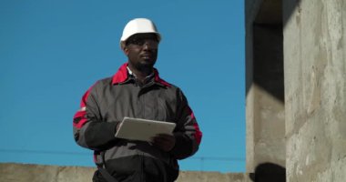 Beyaz şapkalı Afrikalı Amerikalı müteahhit inşaat alanında duruyor ve tabletine notlar yazıyor. Sinema 4K 60fps video