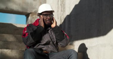 İnşaat alanının beton merdivenlerinde oturan Afrikalı Amerikalı işçiler. Kendini iyi hissetmiyor ve başı ağrıyor. Gözlüklerini çıkarıp kafasına masaj yapıyor. Sinema 4K 60fps video