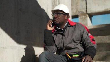 Afrikalı Amerikalı işçiler akıllı telefondan konuşur. Beyaz şapkalı ve koruyucu gözlüklü inşaatçı inşaat alanındaki beton merdivenlerde oturuyor ve cep telefonuyla konuşuyor.
