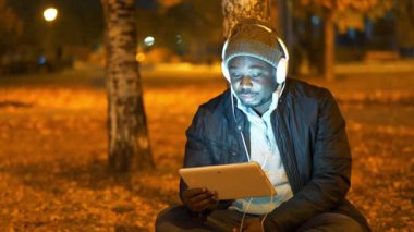 Kışlık şapkalı ve kulaklıklı Afro-Amerikan bir adam akşam parkında oturup tablet bilgisayarla video bağlantısıyla konuşuyor. Yapay ışıklandırmanın açık hava lambalarından titreyen video