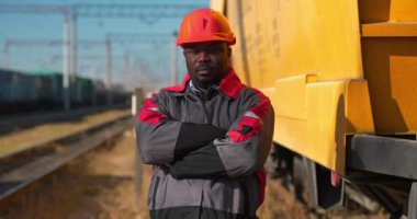 Afrikalı Amerikan demiryolu işçisi yük treni terminalinde duruyor. Demiryolu işçisi üniformalı ve kırmızı başlıklı adam kameraya bakıyor. Sinema 4K 60fps video