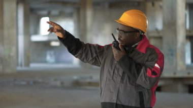 Siyah şapkalı ve gözlüklü Afrikalı Amerikalı işçiler proje sahasında duruyor ve taşınabilir radyo vericisiyle konuşuyor.