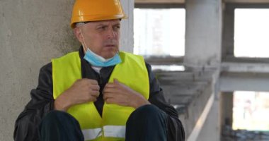 Tıbbi koruyucu yüz maskesi takan işçiler inşaat alanında oturuyor. Kendini iyi hissetmiyor, burnunu siliyor, öksürüyor ve burnunu mendille siliyor.
