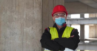 İnşaat alanında mavi tıbbi koruyucu maskeli, gözlüklü ve kırmızı başlıklı adam kameraya bakıyor. Sinema 4K 60fps video