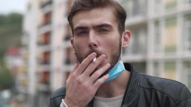 Tıbbi koruyucu maskeli genç adam çenesinde sigara içiyor ve gülümsüyor.
