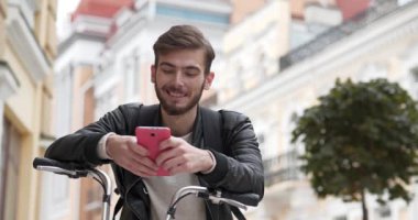 Bisikletli genç yakışıklı adam sokakta duruyor, akıllı telefon kullanıyor ve gülümsüyor. Sırt çantalı bisikletli kurye akıllı telefonuna bakıyor. Sinema 4K 60fps video