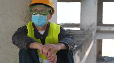 Gözlüklü, sarı şapkalı ve mavi koruyucu maskeli işçiler inşaat alanında oturup etrafa bakınıyorlar. 4K 60fps eğik video