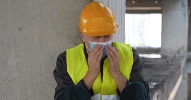 İnşaat alanında mavi tıbbi koruyucu maske ve sarı başlık takan işçiler oturuyor. Kendini iyi hissetmiyor, hapşırıyor, burnunu siliyor, öksürüyor ve burnunu mendille siliyor.
