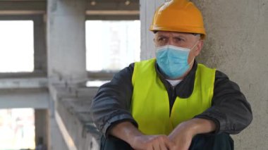 Tıbbi koruyucu yüz maskesi ve başlık takan kişi inşaat alanında oturuyor. Öksürüyor ve iyi hissetmiyor. Göğüs ağrısı var.