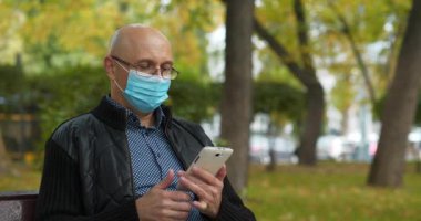 Tıbbi koruyucu maskeli orta yaşlı kel bir adam parktaki bankta oturur, telefon numarasını çevirir ve akıllı telefondan konuşur. Sinema 4K 60fps video