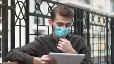 Slow Motion Genç yakışıklı tıbbi koruyucu yüz maskeli adam bankta oturuyor ve tablet bilgisayarla çalışıyor.