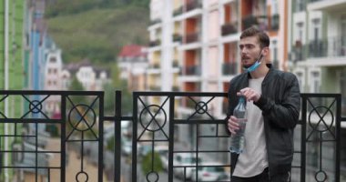 Genç yakışıklı adam tıbbi koruyucu yüz maskesini çıkarıyor ve büyük plastik şişeden maden suyu içiyor. Sinema 4K panoramik yavaş çekim videosu