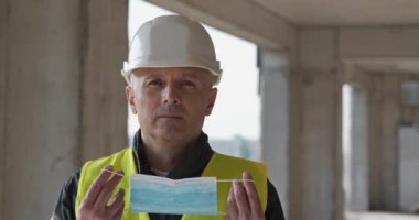 İnşaat alanında çalışan işçi kameraya bakar ve tıbbi koruyucu yüz maskesi takar. Sinema 4K 60fps video