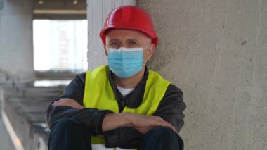 Tıbbi koruyucu yüz maskesi ve kırmızı başlıklı inşaat işçisi yere oturup kameraya bakıyor.