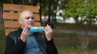 Kel kıdemli adam şehir parkında bankta oturur ve tıbbi koruyucu yüz maskesi takar.