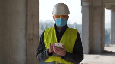 Beyaz başlıklı, turuncu gözlüklü ve mavi tıbbi koruyucu maskeli adam inşaat alanında duruyor, etrafa bakıyor ve akıllı telefonlara veri giriyor.