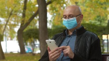 Mavi tıbbi koruyucu maskeli kel bir adam parktaki bankta oturur ve akıllı telefonundaki fotoğraflara bakar, arka planda trafik vardır.
