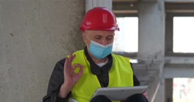 Kırmızı başlıklı ve mavi tıbbi koruyucu maskeli işçiler inşaat alanında oturup tablet bilgisayarla video bağlantısıyla iletişim kuruyor. Sinema 4K 60fps video