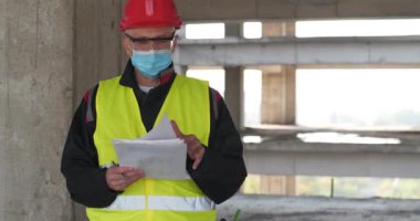 İnşaat işçisi kırmızı şapka ve tıbbi koruyucu maske takıyor inşaat sahasında duruyor, el ele tutuşup çalışma evraklarını tutuyor, etrafa bakıyor ve notlar alıyor. Sinema 4K 60fps video