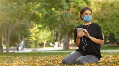 Tıbbi koruyucu maskeli genç bir kız sonbahar parkında çimlerde oturur, akıllı telefonuna bakar ve müzik dinler. Sonra maskesini çıkarıp temiz hava soluyor.