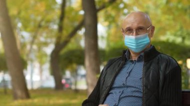 Slow Motion Kel adam mavi tıbbi koruyucu maskeli ve gözlüklü şehir parkındaki bankta oturuyor ve kameraya bakıyor.