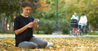 Güz parkında çimenlikte oturan genç bir kız akıllı telefon kullanır, fotoğraflara bakar ve takla atar. Arka planda bebek ve bebek taşıyıcısı olan insanlar yürüyor. Sinema 4K 60fps video