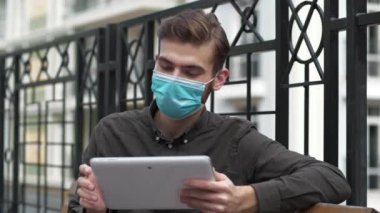 Tıbbi koruyucu maskeli genç adam bankta oturur ve tablet bilgisayar kullanır, fotoğraf çeker ve bakar.