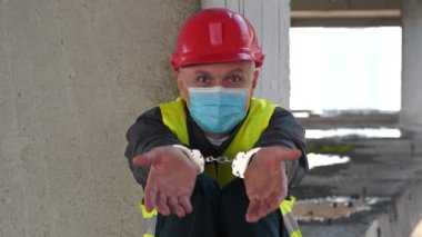 Kırmızı başlıklı ve tıbbi koruyucu maskeli Yavaş Hareketli Kelepçeli işçi inşaat alanının beton zemininde oturuyor. Kameraya bakar, konuşur ve el kol hareketi yapar.