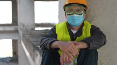 Gözlüklü, kasklı ve tıbbi koruyucu maskeli işçiler inşaat alanında oturur ve kameraya bakar. 4K 60fps eğik video