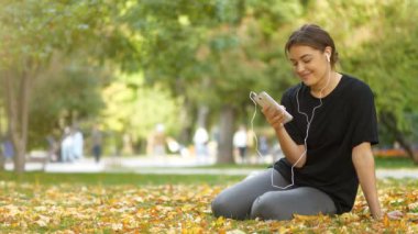 Genç, güzel bir kız, sonbahar parkında sarı yaprakların üzerinde oturur, akıllı telefona bakar ve müzik dinler.