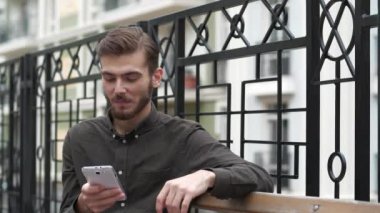 Adam bankta oturuyor ve akıllı telefonundaki fotoğraflara bakıyor, gülümsüyor ve keyfi yerinde.