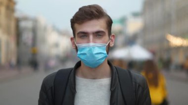 Koruyucu maskeli ve sırt çantalı bir adam kalabalık caddede dikilip kameraya bakıyor. 4K Geri Zaman Yavaş Hareket videosu