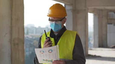 Tıbbi koruyucu maske, gözlük ve başlık takan işçiler inşaat sahasında duruyor, el işi belgelerini tutuyor ve taşınabilir radyo vericisiyle konuşuyor.