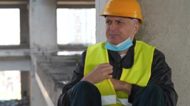 Mavi tıbbi koruyucu maskeli ve sarı başlıklı Yavaş Hareket İşçisi inşaat alanında oturuyor. Kendini iyi hissetmiyor, maskesini çıkarıyor, hapşırıyor, burnunu siliyor ve tekrar yüz maskesi takıyor.