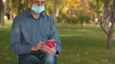 Tıbbi koruyucu maskeli kıdemli adam parkta bankta oturuyor ve akıllı telefon kullanıyor. Covid konsept sağlık ve güvenlik, koronavirüs karantinası, sonraki dalga covid virüs koruması