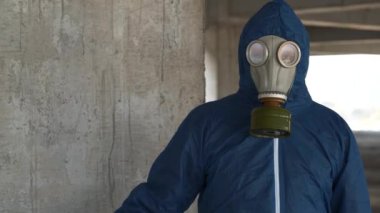 Gaz maskeli ve koruyucu giysili bir adam endüstriyel alanda duruyor, elinde ölü bir kuş var ve kameraya bakıyor.